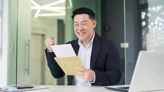 微笑的亚洲商人坐在办公室的办公桌前读着带来好消息的信