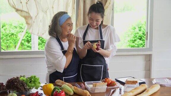 快乐的亚洲小女儿和年长的母亲在平板电脑上烹饪在线课程一起在家里的厨房里做新鲜的蔬菜食物