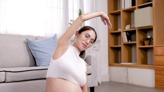 一名怀孕的亚洲少女妈妈穿着短上衣露出她日渐隆起的腹部坐在自家客厅的地板上做瑜伽姿势同时摇晃着身体放松期待着自己的分娩