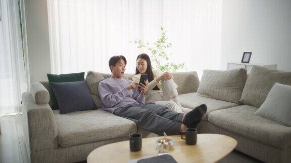 不同的韩国夫妇在明亮舒适的客厅沙发上玩智能手机一起享受时光寻找度假目的地的青年男女