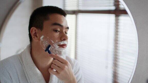 亚洲男子站在浴室镜子旁用剃须刀刮胡子