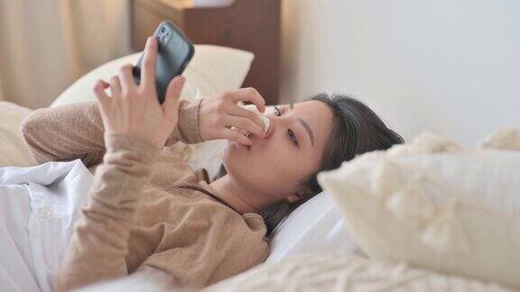 一名亚洲华裔妇女躺在床上盖着毯子患感冒和流感用智能手机给朋友发短信用数字温度计测量体温