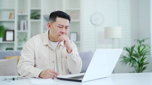 失望的亚洲人坐在家里办公室的办公桌前在笔记本电脑上看坏消息