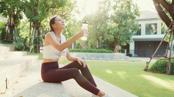美丽年轻的亚洲运动员女士锻炼喝水因为在城市公园环境中跑步后感到累在晚上