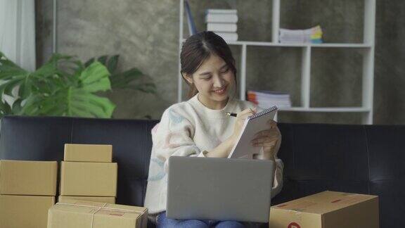 亚洲妇女坐在沙发上坐在家里的盒子和笔记本电脑从客户网上业务中小企业销售概念