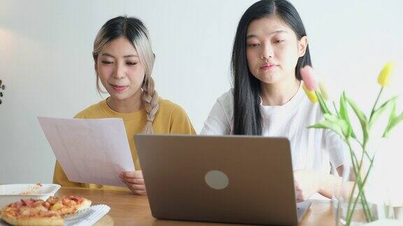 两位年轻的亚洲女性朋友愉快地吃着披萨、沙拉和意大利面一边聊天一边在家里用笔记本电脑工作亚洲休闲餐厅