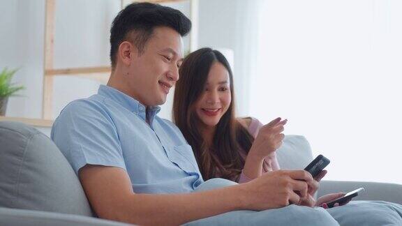 年轻幸福的亚洲夫妇坐在沙发上放松在客厅亚洲夫妇男人和女人喜欢使用智能手机连接互联网看社交媒体花时间在家里