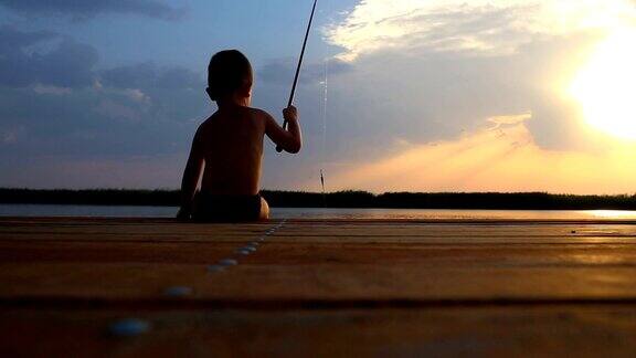 后视图的小男孩坐在一个木制码头的边缘和钓鱼的湖在日落