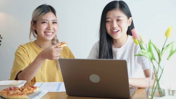 两位年轻的亚洲女性朋友愉快地吃着披萨、沙拉和意大利面一边聊天一边在家里用笔记本电脑工作亚洲休闲餐厅