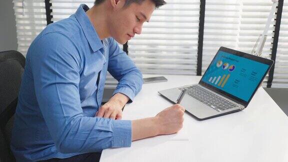亚洲商人用笔记本电脑分析商业营销帅气的职业男员工坐在桌子上开心地在纸上写纸条在工作场所工作