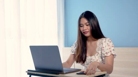 4K50fps美丽的亚洲女孩穿着花衬衫正兴高采烈地在笔记本电脑上订购手里拿着信用卡拿起信用卡付款网上购物