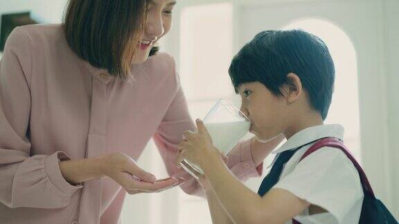 美丽的亚洲妈妈正在鼓励穿着校服和背包的小儿子在早上上学前喝鲜奶以保持孩子健康的生活方式小男孩在家用玻璃杯喝鲜奶