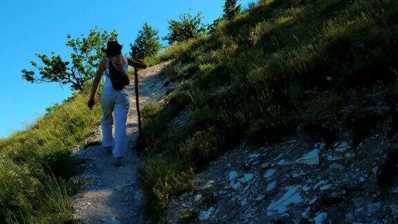 一个女孩背着双肩包拄着一根木棍走在山间陡峭山坡上的小路上