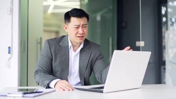 在现代办公室里穿着正装的沮丧的亚洲人坐在办公室的笔记本电脑上看坏消息