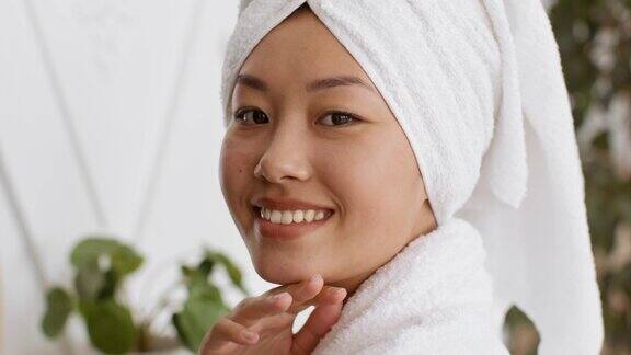 健康愉快的亚洲妇女头戴毛巾对着镜头微笑在脸上涂抹保湿霜