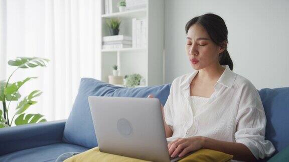 年轻的亚洲女性坐在家里客厅的沙发上用笔记本电脑与顾客进行视频通话