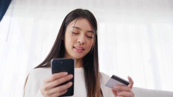 年轻的亚洲女子坐在客厅沙发上用智能手机上的银行卡通过互联网进行网上银行支付用信用卡在手机上网上购物