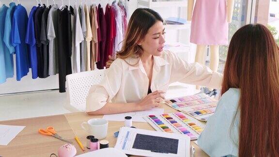 两位亚洲女性时装设计师或裁缝集思广益讨论新系列的设计并在平板电脑上展示面料样品和草图在演播室一起工作