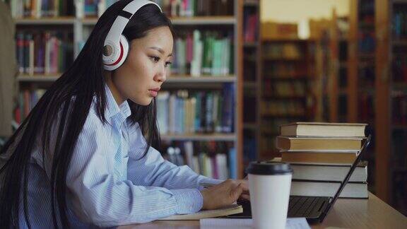 美丽的亚洲女学生戴着耳机坐在图书馆的书桌前专注于学校项目年轻女孩在笔记本电脑上打字喝咖啡在抄写本上写字