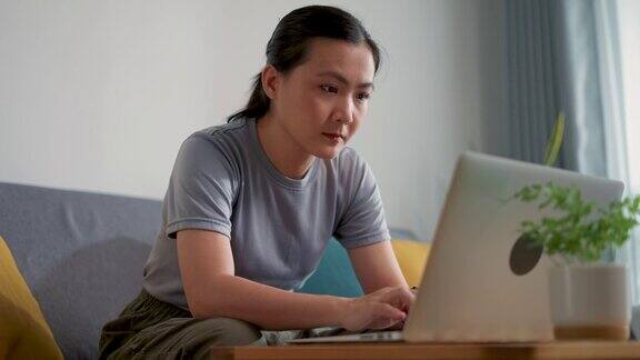 亚洲女性坐在家里的客厅里用笔记本电脑和键盘打字