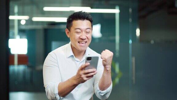 快乐的亚洲人员工享受成功在智能手机上阅读好消息商人赢得手机在线投注出价金钱奖金获胜收到提供机会的智能手机