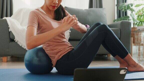 年轻迷人的亚洲女性在运动服装观看健身在线视频平板电脑与健身球在客厅的房子