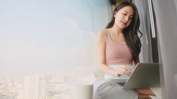 幸福迷人的亚洲女性放松休闲坐在窗边看外面的手使用笔记本电脑在家里工作