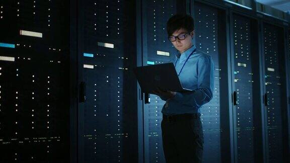在黑暗的数据中心:男性IT专家走在一排操作服务器机架上使用笔记本电脑进行维护云计算、人工智能、超级计算机、网络安全