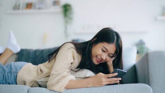 快乐的年轻亚洲女人在家里舒适的沙发上放松地用智能手机发短信微笑的女孩用手机在小工具上浏览无线互联网在家网上购物