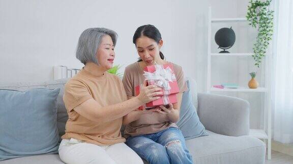 亚洲美女女儿惊喜她的母亲与生日礼物年轻漂亮的女孩坐在家里的沙发上在客厅给年长的老妈妈送礼盒祝贺