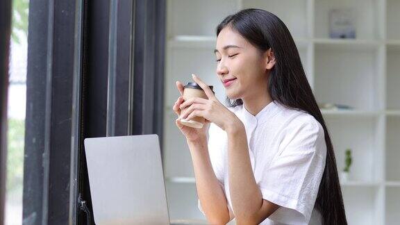 快乐的亚洲女性在笔记本电脑上工作在舒适的家里喝咖啡