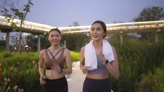前视图:两个亚洲美丽的中年女性朋友在运动服装完美的肌肉一起积极慢跑在黄昏时间使用智能手表监测在行人天桥背景的人行道在公园花园积极的生活方式