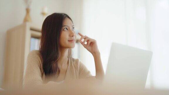 年轻的亚洲妇女在家里客厅桌上的笔记本电脑上工作