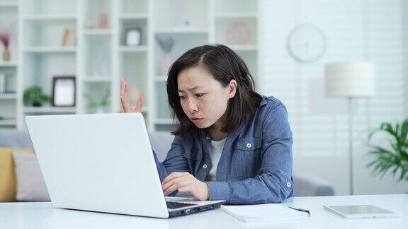 失望的亚洲女人坐在家里的办公室里在笔记本电脑上读坏消息