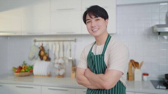 快乐的年轻亚洲男子系着围裙双臂交叉站在家里的厨房里对着镜头微笑
