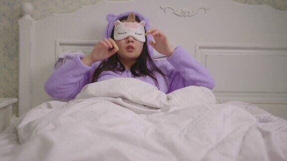 20多岁的亚洲女性戴着睡眠面具躺在床上穿着紫色睡衣的女人打着哈欠戴着睡眠面罩躺在枕头上