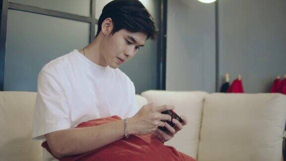 一名年轻的亚洲男子坐在家里的沙发上用智能手机玩电子游戏