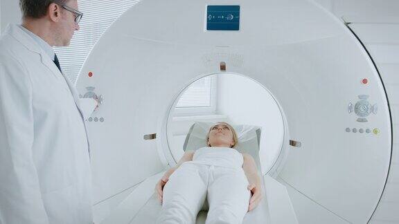 在医学实验室放射学家控制MRI或CT或PET扫描与女性病人正在进行的程序高科技现代医疗设备摄像头安装在床上随病人移动