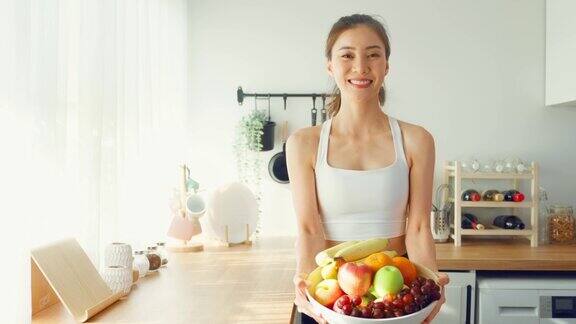亚洲有吸引力的女人的肖像持有水果碗看着相机美丽的运动女孩在运动服享受吃干净的蔬菜锻炼后的健康在家里饮食与健康食品理念