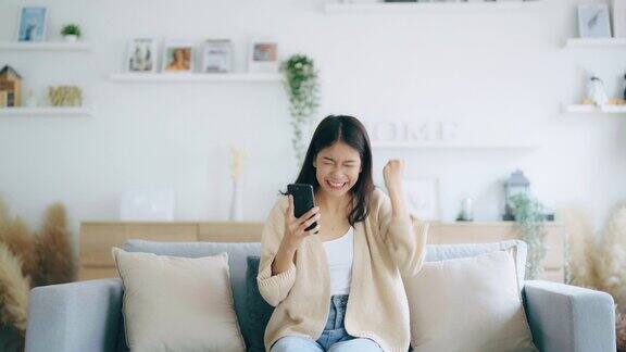 快乐的年轻亚洲女人放松在家里舒适的沙发上发短信在智能手机上微笑的女孩使用手机聊天在线消息从家里网上购物