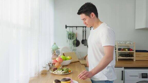 亚洲活跃的帅哥拒绝吃甜甜圈喜欢吃健康食品年轻有魅力的男性在厨房的碗里吃蔬菜沙拉在桌子上喝干净的水饮食与健康理念