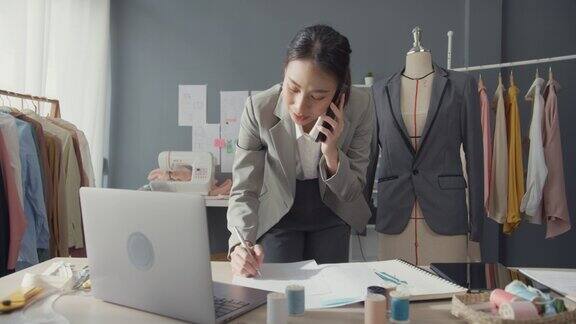 专业开朗的亚洲女商人设计师坐在办公桌上用笔记本电脑打电话给客户在工作室工作场所的网上市场确认购买订单成功的事业