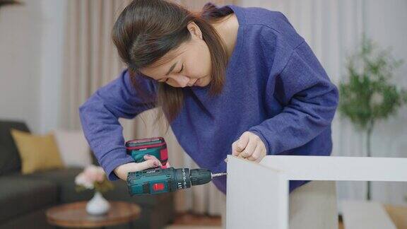年轻的亚洲妇女在家里自己用电钻组装新家具一名妇女自己用钻头拧紧家具组装上的螺丝拼装家具概念