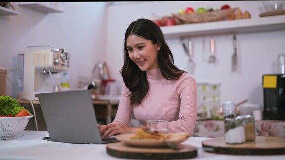 微笑的年轻亚洲女子坐在厨房里在家里的厨房里用笔记本电脑看搜索网上博客在家里享受周末