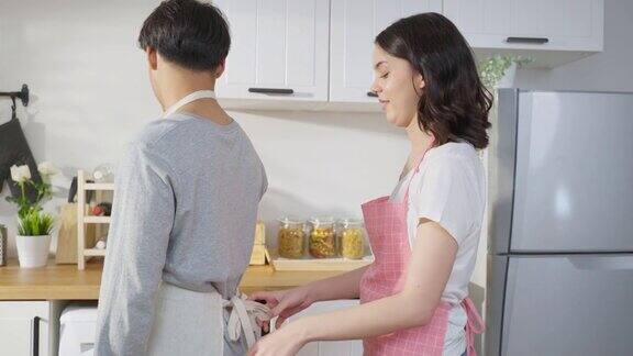 亚洲年轻新婚妻子在厨房给丈夫系围裙鞋带有魅力的浪漫情侣穿着围裙在家里一起度过时光心情愉快享受做饭、烘焙、烘焙的乐趣