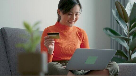 亚洲女性手持信用卡用笔记本电脑在家里的客厅里网上购物