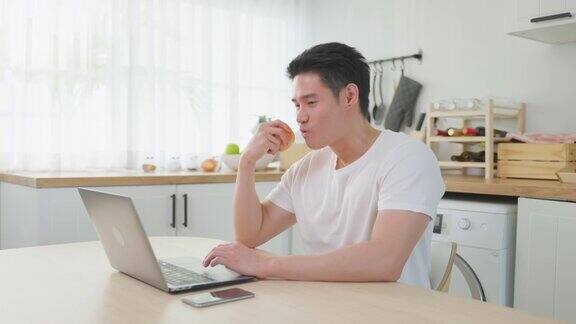 亚洲商人在家工作时吃苹果健康食品有吸引力的英俊年轻男性感觉快乐和享受使用笔记本电脑学习在线学习和吃水果在家里的厨房