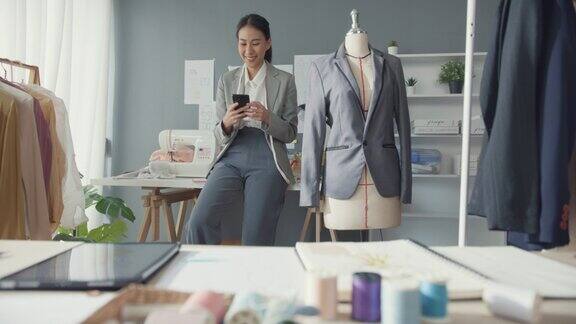 开朗的亚洲女商人时装设计师与套装系列微笑坐在桌子上用智能手机与客户聊天确认在线采购订单在工作室工作场所开始网上业务