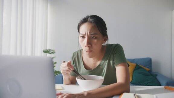 亚洲商务女性穿着休闲吃方便面在家里客厅用笔记本电脑工作