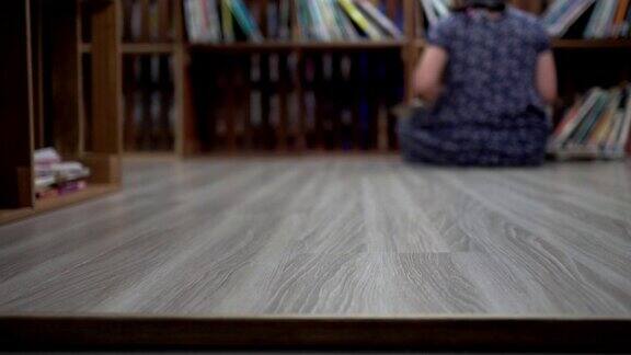 小女孩坐在木地板上看书以下的脖子背对着镜头在那个女孩的旁边有一个书架焦点在前景
